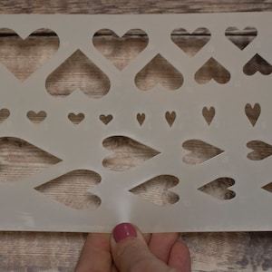 Puede incluir: Una plantilla de plástico blanco con múltiples formas de corazón de diferentes tamaños. La plantilla está diseñada para manualidades y la creación de diseños con forma de corazón.