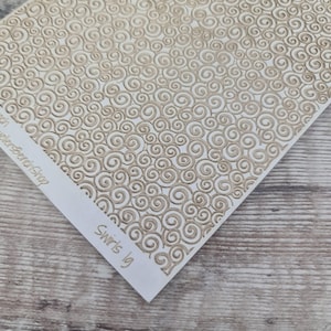 May include: A white paper featuring a repeating pattern of beige swirls. The paper is angled, with a corner displaying the text "Swirls 1g". The background is a light wood grain.