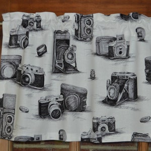 May include: A white valance with a black and white pattern of vintage cameras. The valance has a black and white checkered pattern and features various camera designs.