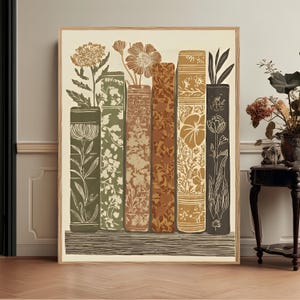 Op de afbeelding: Een rechthoekige wandkunstprint in een lichthouten lijst met een verticale illustratie van zes boeken op een plank. De ruggen van de boeken zijn versierd met diverse bloem- en plantmotieven in tinten olijfgroen, beige en bruin.