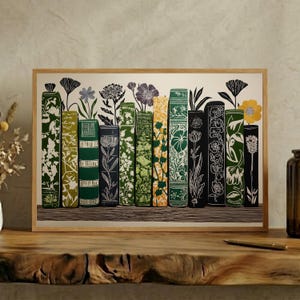 May include: A rectangular framed art print featuring a row of eleven book spines standing upright. Each spine is decorated with intricate botanical illustrations in shades of green, black, and yellow, with flowers extending from the top of the books.