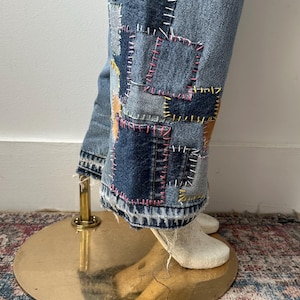 May include: Light blue denim jeans with patchwork detail on the lower legs. Patches are various shades of blue and tan, stitched with pink, yellow, and white thread. The jeans are displayed on a mannequin.