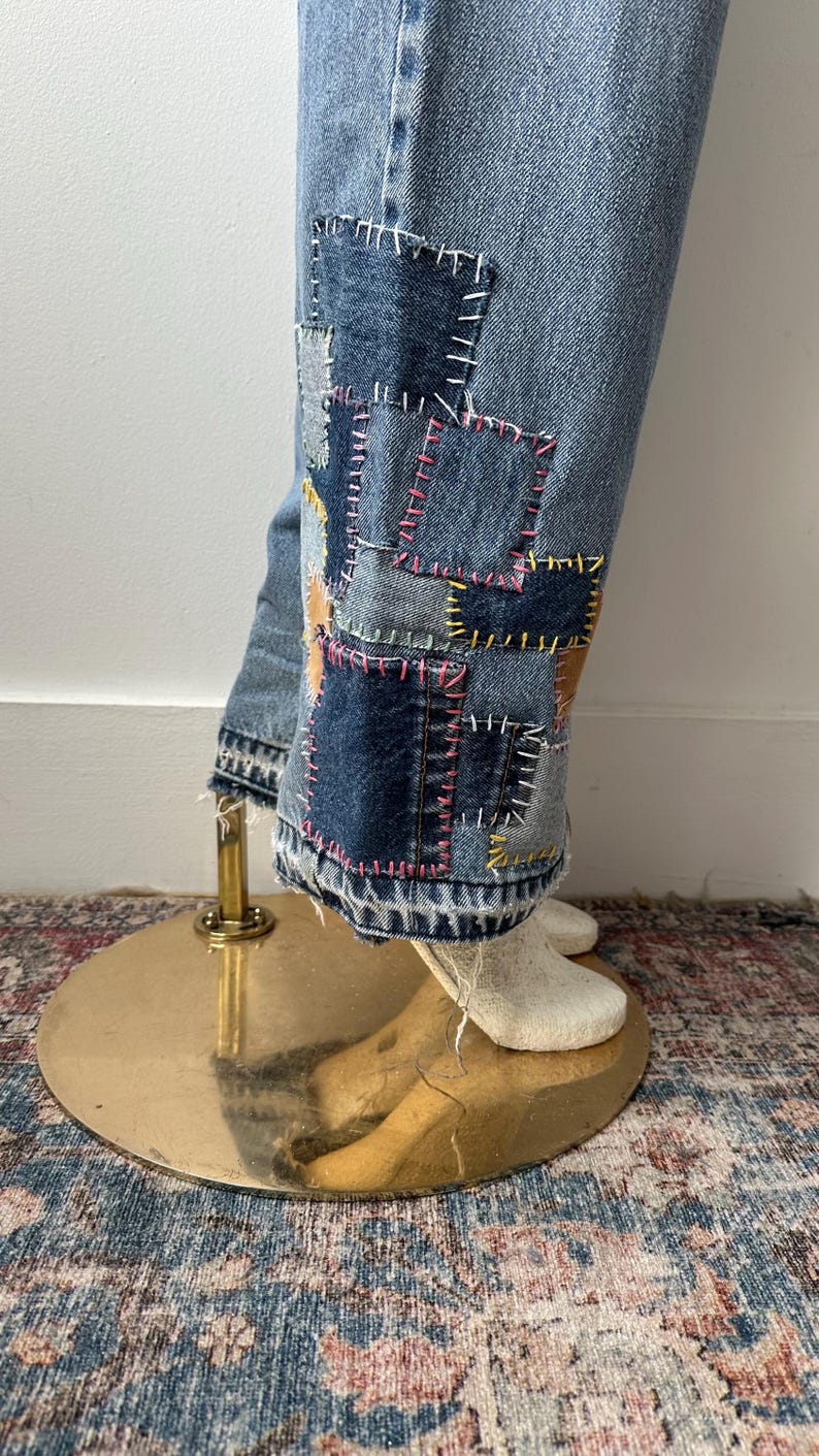 May include: Light blue denim jeans with patchwork detail on the lower legs. Patches are various shades of blue and tan, stitched with pink, yellow, and white thread. The jeans are displayed on a mannequin.