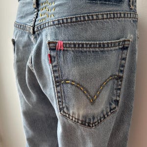 May include: Light blue denim jeans with yellow stitching and a red tag on the back pocket. The jeans have a distressed look with a faded wash. The back pocket has a pink accent.