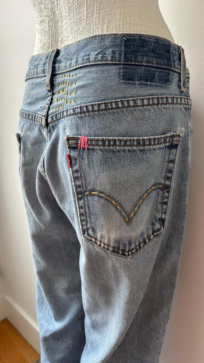 May include: Light blue denim jeans with yellow stitching and a red tag on the back pocket. The jeans have a distressed look with a faded wash. The back pocket has a pink accent.