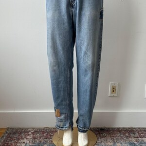 May include: Light blue denim jeans with a relaxed fit, featuring patches on the knees and cuffs. The jeans are displayed on a mannequin, showcasing their vintage style. The jeans have a classic five-pocket design.
