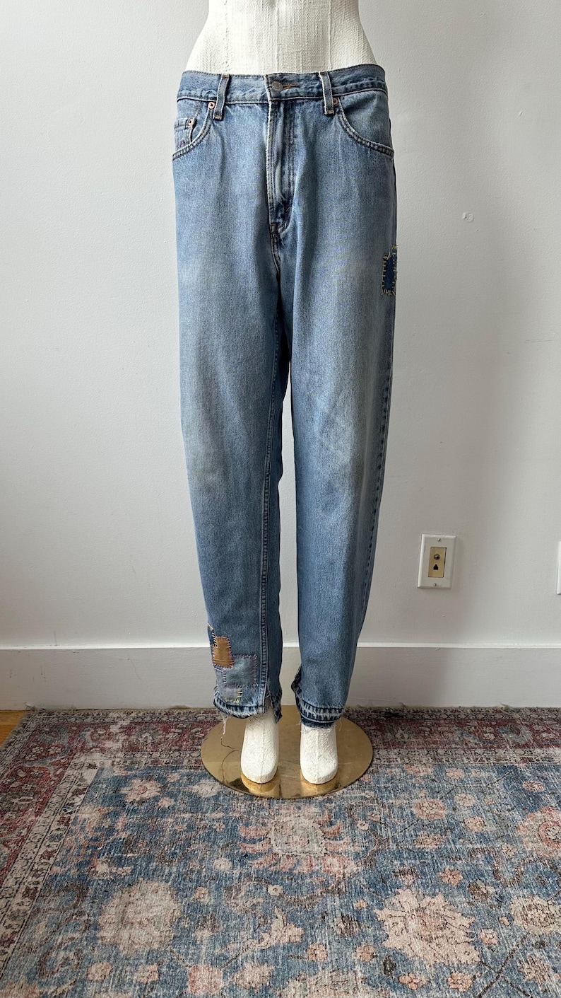 May include: Light blue denim jeans with a relaxed fit, featuring patches on the knees and cuffs. The jeans are displayed on a mannequin, showcasing their vintage style. The jeans have a classic five-pocket design.