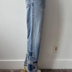 May include: Light blue denim jeans with patchwork detail at the ankles. The jeans are displayed on a mannequin. The patchwork features various shades of blue and purple. The jeans have a classic five-pocket design.