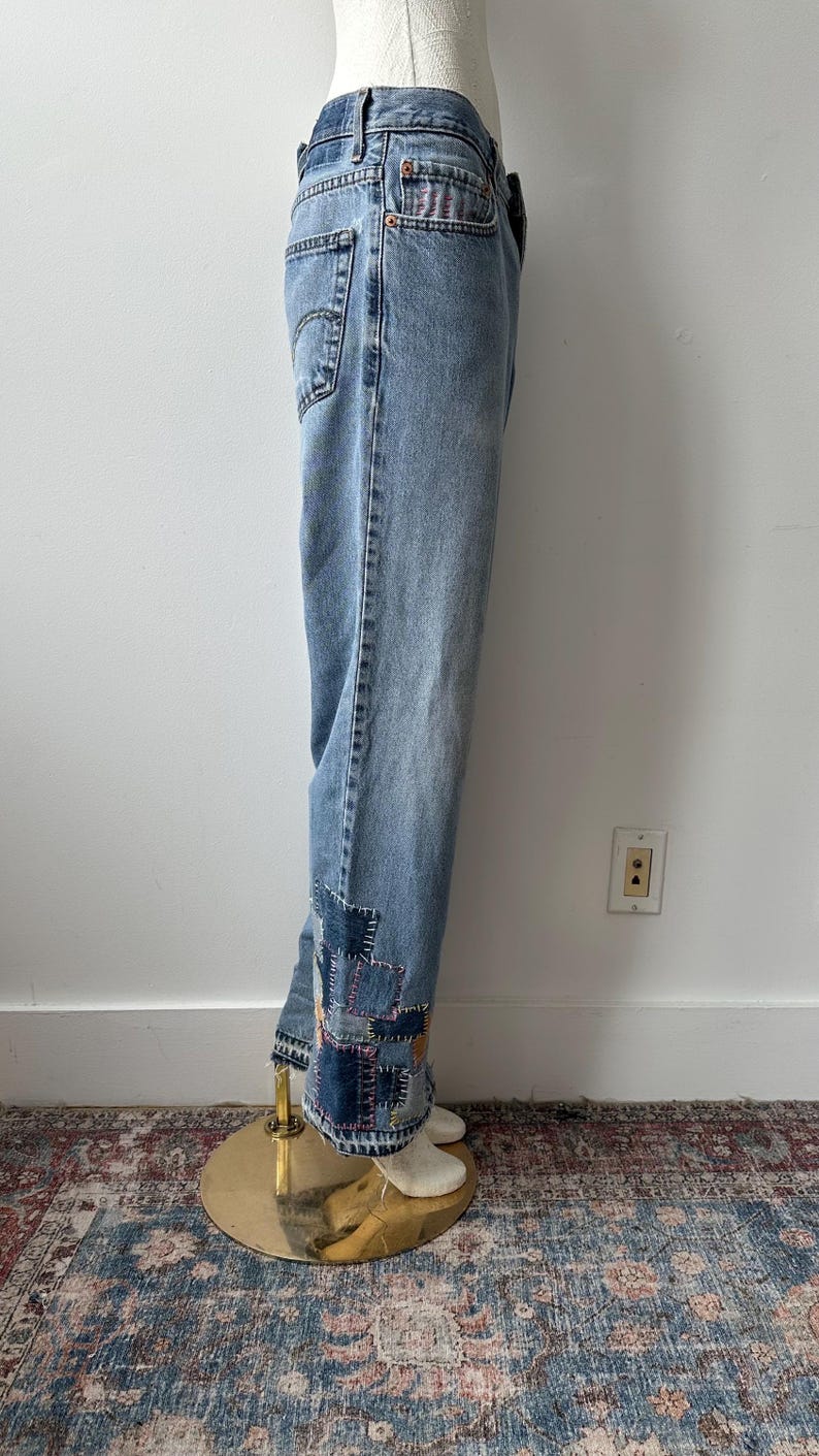 May include: Light blue denim jeans with patchwork detail at the ankles. The jeans are displayed on a mannequin. The patchwork features various shades of blue and purple. The jeans have a classic five-pocket design.