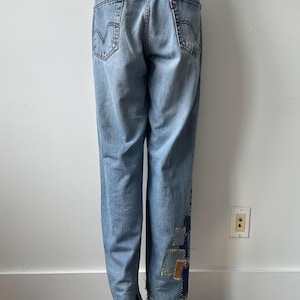 May include: Light blue denim jeans with patches on the lower leg. The jeans have a classic five-pocket design, a red Levi's tag, and a light wash. The jeans are displayed on a mannequin.
