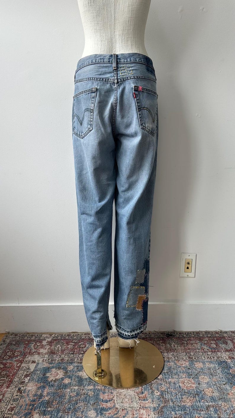 May include: Light blue denim jeans with patches on the lower leg. The jeans have a classic five-pocket design, a red Levi's tag, and a light wash. The jeans are displayed on a mannequin.