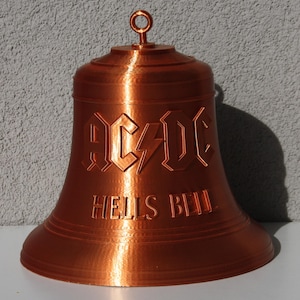 Könnte beinhalten: Eine kupferfarbene Glocke mit dem geprägten Schriftzug AC/DC und HELLS BELL auf der Vorderseite. Die Glocke hat eine glatte, ausgestellte Basis und eine kleine kreisförmige Öse an der Oberseite zum Aufhängen, mit metallischem Glanz und sichtbaren Schichten.
