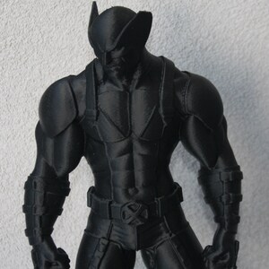 Peut inclure: Une figurine noire mate imprimée en 3D représentant un personnage musclé portant un masque aux oreilles pointues, une combinaison tactique avec des bretelles et une ceinture ornée d'un emblème en forme de X. Les bras sont musclés et des griffes dépassent.