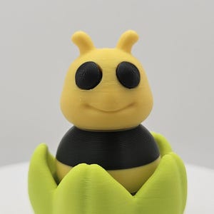 May include: A 3D-printed yellow and black bee figurine with large black eyes, perched inside a light green flower-like base. The bee has a smiling expression and small antennae. The figurine is made of plastic.