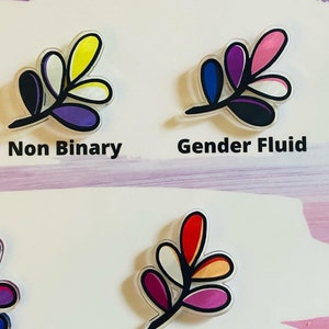 May include: Four different colored leaf designs with black outlines. The leaves are in the colors of the non-binary, gender fluid, lesbian, and bisexual pride flags. The text "Non Binary" and "Gender Fluid" are written below the first two designs.