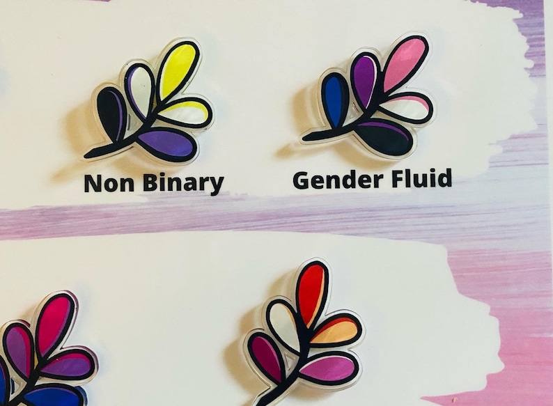 May include: Four different colored leaf designs with black outlines. The leaves are in the colors of the non-binary, gender fluid, lesbian, and bisexual pride flags. The text "Non Binary" and "Gender Fluid" are written below the first two designs.