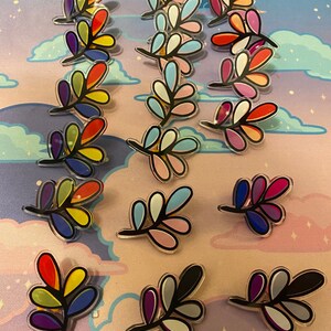May include: A collection of 15 colorful leaf-shaped pins. Each pin features a different color scheme, including rainbow, blue, pink, and black.