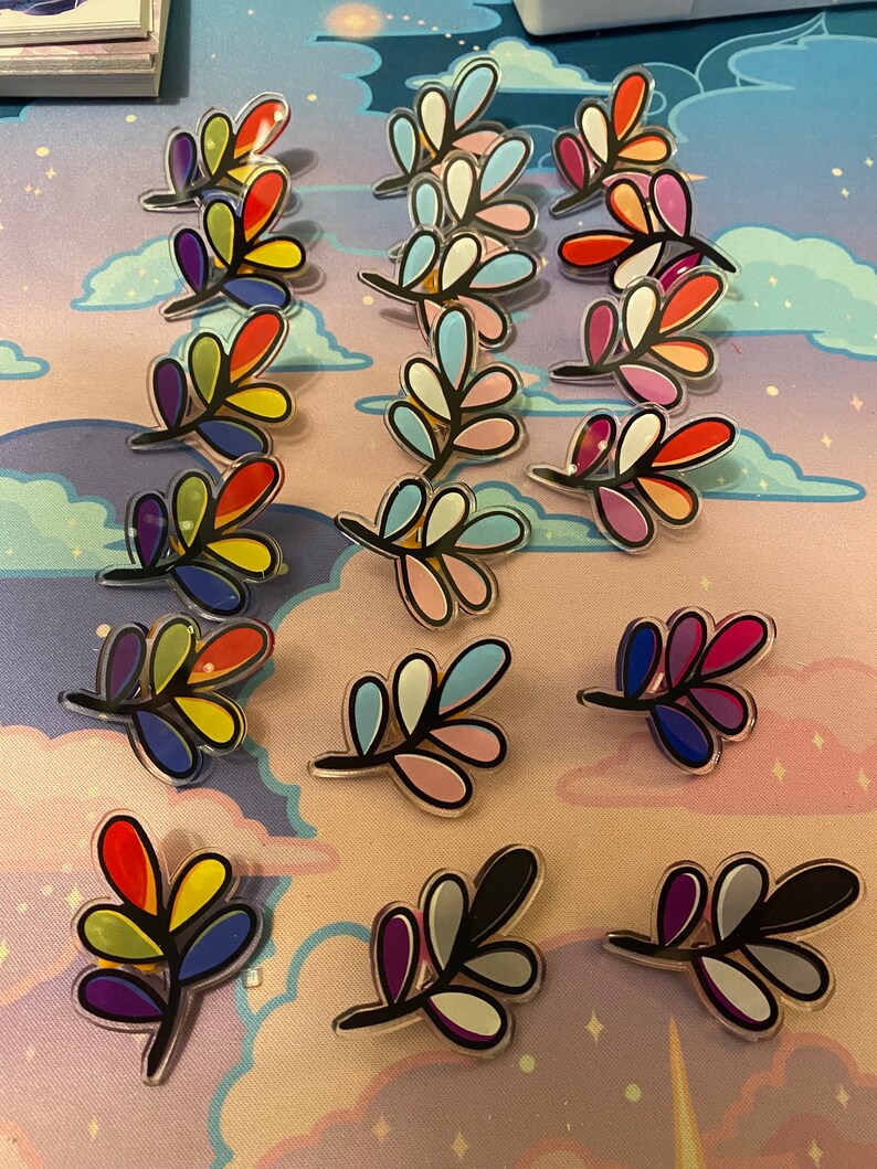 May include: A collection of 15 colorful leaf-shaped pins. Each pin features a different color scheme, including rainbow, blue, pink, and black.