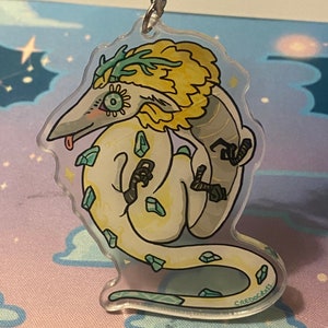 May include: A white and yellow dragon with green antlers and a light blue tongue. The dragon is surrounded by light blue crystals and has a yellow mane. The dragon is on a clear acrylic keychain with the text "CREDOCAST" on the bottom.
