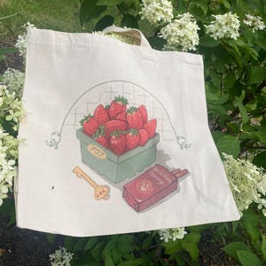 May include: A cream-colored canvas tote bag featuring a graphic print of a green basket filled with red strawberries, a gold-colored key labeled 707, and a red rectangular box labeled Black Stone Cherry, all framed by a thin green arched line.