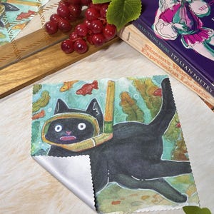 May include: A square microfiber cleaning cloth featuring a watercolor illustration of a black cat wearing a yellow snorkel and mask, swimming underwater among green seaweed and small orange fish. The cloth has a white back and scalloped edges.