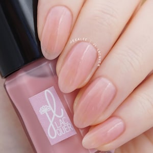 May include: A bottle of pink nail polish with a black cap is held in a hand. The nails are painted with a matching pink polish. The label on the bottle has a floral design and the text "JL Lacquer". The text "2 coats absolut" is visible.