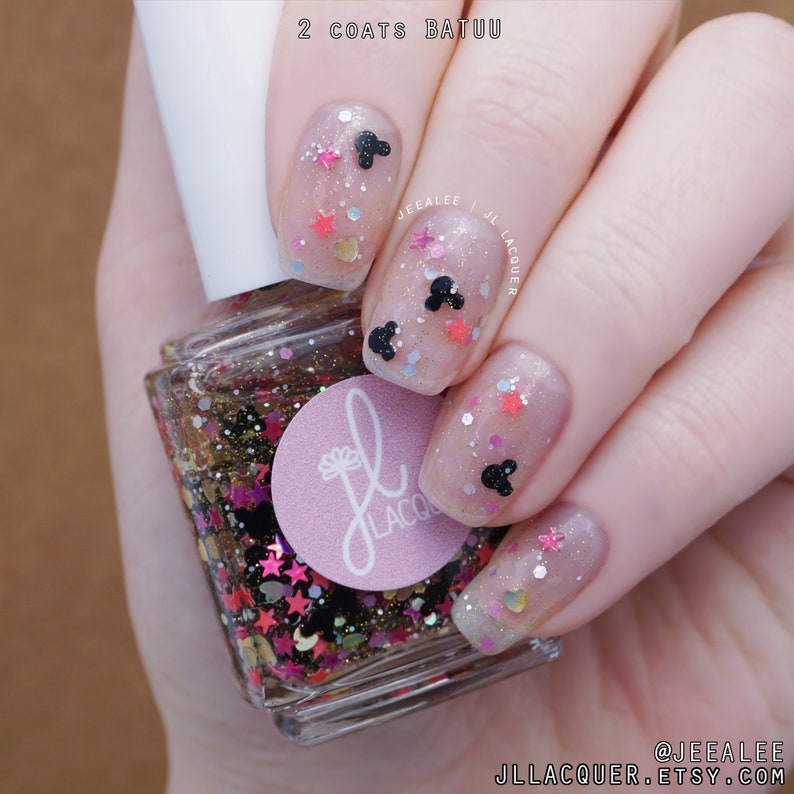 May include: A clear nail polish bottle with a pink label that says "JL LACQUER" and a black and white Mickey Mouse head design. The bottle is surrounded by glitter and stars. The image shows a hand with nails painted with the polish, featuring Mickey Mouse heads and stars.