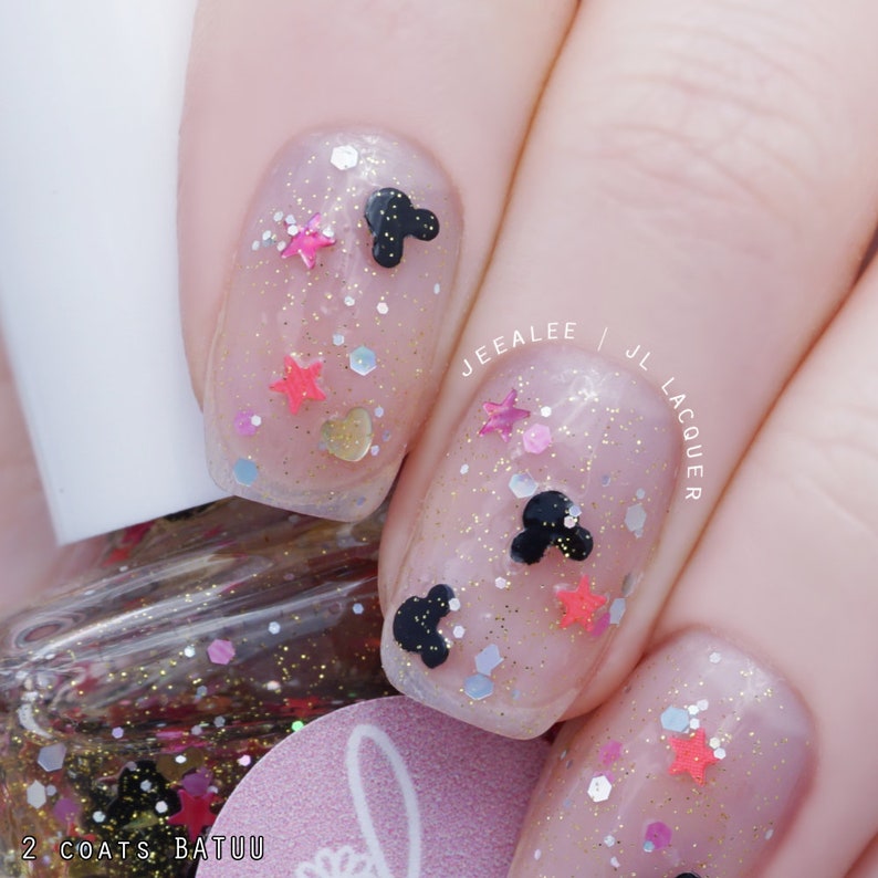 May include: Close-up of fingernails with a clear base coat and glitter, featuring black Mickey Mouse heads and pink, red, and gold stars. The text "JEEALEE | JL LACQUER" is visible on the right fingernail. The bottle of nail polish is labeled "2 COATS BATUU".
