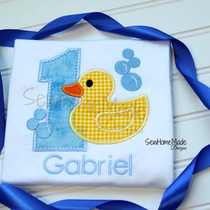 May include: White shirt with a blue number 1 and a yellow gingham duck with an orange beak. The shirt has the name "Gabriel" embroidered in blue. Blue bubbles and a blue ribbon frame the shirt.