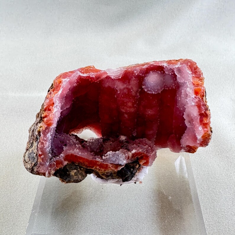 Puede incluir: Una geoda abierta y vibrante con un interior rojo intenso y un revestimiento cristalino rosa-p&uacute;rpura m&aacute;s claro. La c&aacute;scara exterior es una mezcla de tonos marrones y anaranjados, creando un contraste llamativo. La geoda se exhibe sobre un soporte transparente.