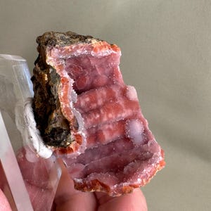 Puede incluir: Un esp&eacute;cimen mineral natural con una vibrante estructura cristalina rosa y roja. La formaci&oacute;n rocosa tiene un exterior rugoso y texturizado y un interior en capas. El esp&eacute;cimen est&aacute; montado sobre una base rectangular transparente.