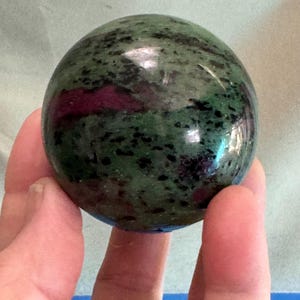 May include: A polished sphere of ruby in zoisite. The stone features a mottled green base with black speckles and patches of deep red. The sphere is held in a hand, showcasing its smooth, reflective surface.