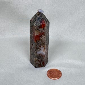May include: A tall, hexagonal-shaped stone obelisk with a polished top and a small, round, light-colored stone on top. The obelisk is primarily dark gray with red and white inclusions. A penny is next to the obelisk for scale.