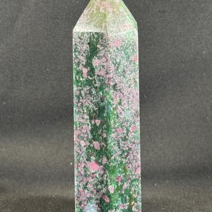 May include: A tall, polished crystal obelisk. The stone features a mix of green and pink hues, with a pointed top. The obelisk is a natural stone, with a textured surface. The crystal is standing upright.
