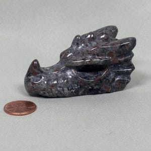 May include: A carved stone dragon head sculpture in shades of gray and maroon. The dragon's features are detailed, with visible scales and a textured surface. A penny is next to the sculpture for size comparison. The sculpture is on a light gray surface.