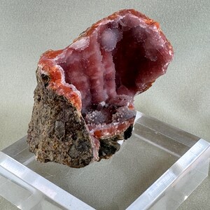 Puede incluir: Un esp&eacute;cimen de geoda natural con un interior hueco revestido de cristales rosa claro brillantes. La capa exterior es de color marr&oacute;n oscuro rugoso con bordes rojo-anaranjados. La geoda se exhibe sobre un soporte acr&iacute;lico transparente.
