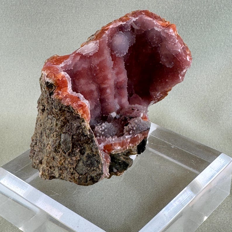 Puede incluir: Un esp&eacute;cimen de geoda natural con un interior hueco revestido de cristales rosa claro brillantes. La capa exterior es de color marr&oacute;n oscuro rugoso con bordes rojo-anaranjados. La geoda se exhibe sobre un soporte acr&iacute;lico transparente.