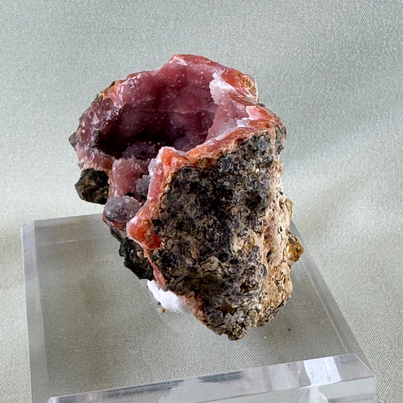 Puede incluir: Esp&eacute;cimen mineral natural con una geoda de interior rosa vibrante. La capa exterior es &aacute;spera, texturizada, marr&oacute;n y negra. La geoda se encuentra sobre un soporte de exhibici&oacute;n acr&iacute;lico transparente, mostrando su estructura cristalina &uacute;nica.