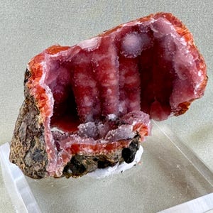 Puede incluir: Un vibrante esp&eacute;cimen mineral natural que presenta una cavidad revestida de formaciones cristalinas. El interior muestra un tono rojo intenso, contrastado por una capa exterior rosa y naranja m&aacute;s clara. La base es una roca oscura y texturizada.