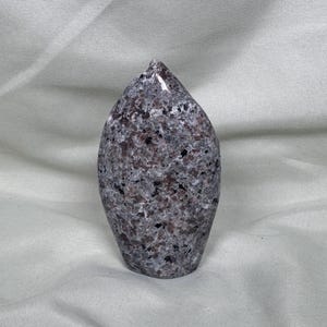 May include: A polished, teardrop-shaped stone sculpture. The stone has a speckled pattern of gray, black, and reddish-brown hues. The sculpture is smooth and glossy, with a pointed top and rounded base. The background is a light gray fabric.