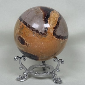 May include: A polished, spherical gemstone with a mix of brown, tan, and gray hues. The orb is displayed on an ornate, silver-colored metal stand. The stone's surface has a natural, patterned appearance.
