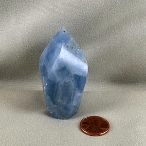 May include: A faceted, light blue crystal point with a rounded base. A copper penny is next to the crystal for size comparison. The crystal has a smooth, polished surface and a slightly irregular shape. The crystal is approximately 3 inches tall.