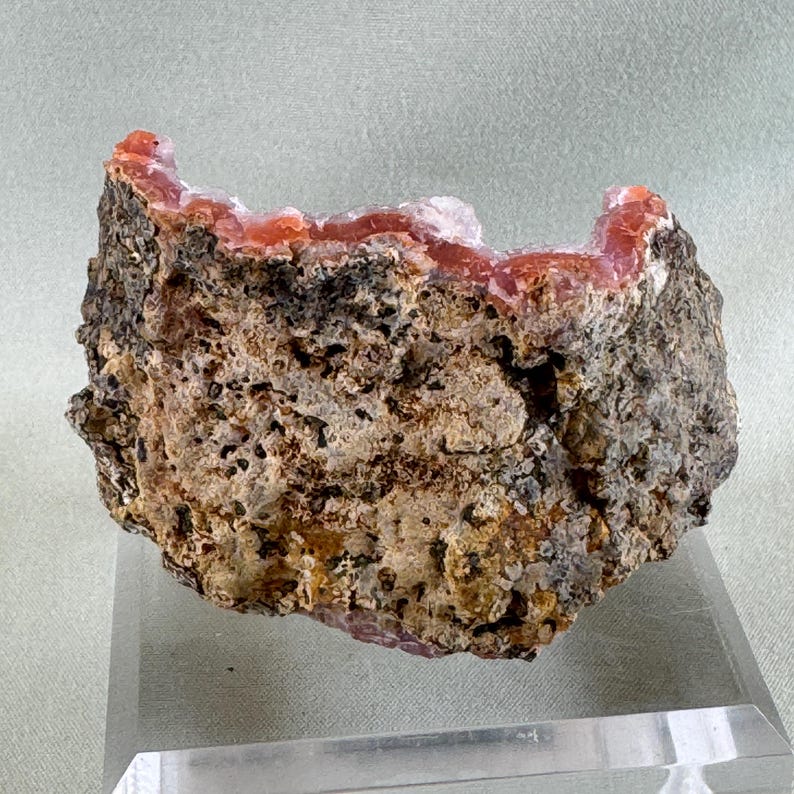 Puede incluir: Una formaci&oacute;n rocosa rugosa y texturizada con un interior cristalino de color coral vibrante. El exterior es una mezcla de gris, marr&oacute;n y negro, creando un contraste llamativo. La muestra se exhibe sobre un soporte rectangular transparente.