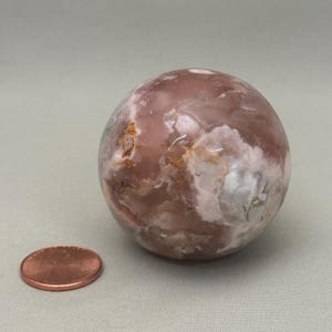 May include: A polished, spherical gemstone with a mix of pink, brown, and white hues. The stone's surface has a marbled appearance. A copper-colored penny is next to the stone for size comparison.