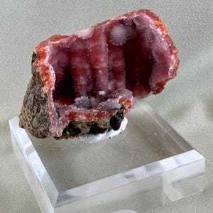 Puede incluir: Un vibrante esp&eacute;cimen mineral rosa con un interior cristalino, exhibido sobre un soporte acr&iacute;lico transparente. La capa exterior presenta tonos marrones y anaranjados, creando un contraste llamativo. El interior presenta una estructura texturizada en forma de cueva.