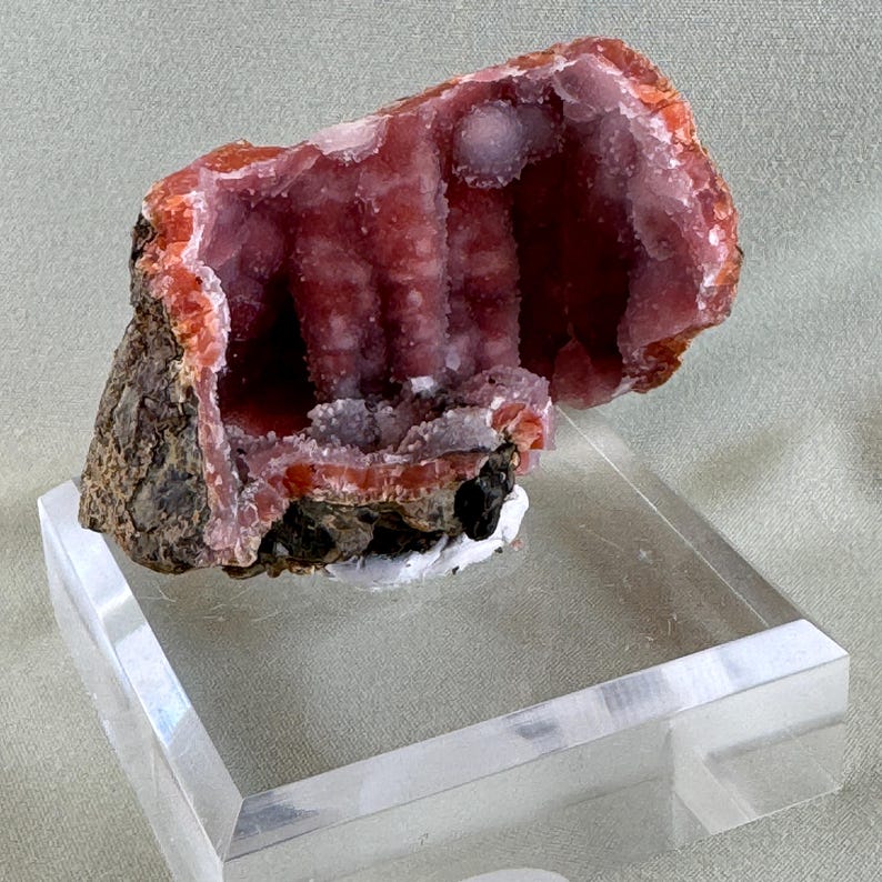 Puede incluir: Un vibrante esp&eacute;cimen mineral rosa con un interior cristalino, exhibido sobre un soporte acr&iacute;lico transparente. La capa exterior presenta tonos marrones y anaranjados, creando un contraste llamativo. El interior presenta una estructura texturizada en forma de cueva.