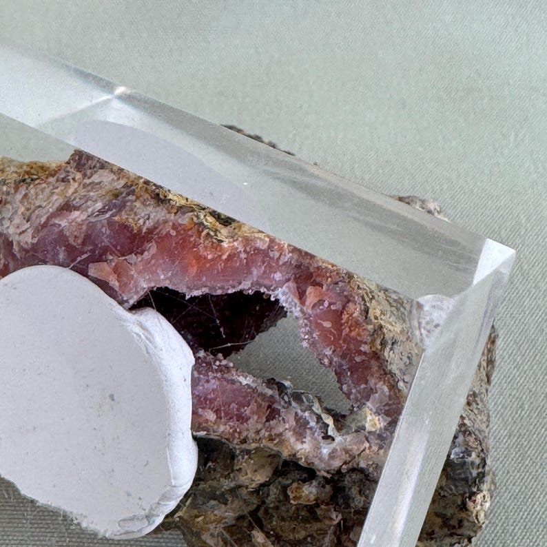 Puede incluir: Un prisma de cristal transparente y rectangular descansa sobre una geoda rugosa de color marr&oacute;n rojizo con un objeto blanco de forma irregular en primer plano. La geoda tiene un centro hueco. El fondo es de color verde claro.