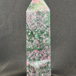 May include: A tall, rectangular crystal obelisk with a pointed top. The stone features a mottled pattern of pink, green, and white. The obelisk is standing upright against a dark background.