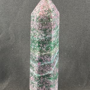 May include: A tall, faceted crystal obelisk. The stone features a mottled pattern of green, pink, and white. The obelisk has a pointed top and a rectangular base. The crystal is a natural stone.