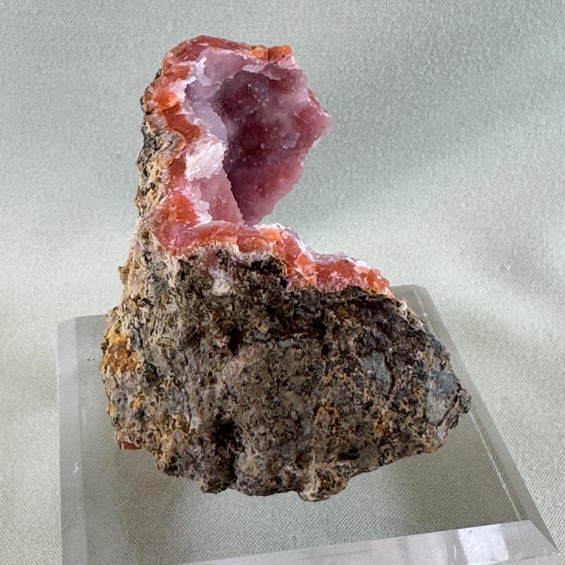 Puede incluir: Un esp&eacute;cimen de geoda con un exterior &aacute;spero de color marr&oacute;n oscuro y un interior rosa vibrante. La cavidad revestida de cristales est&aacute; enmarcada por una banda naranja y blanca. La geoda se exhibe sobre un soporte acr&iacute;lico transparente.